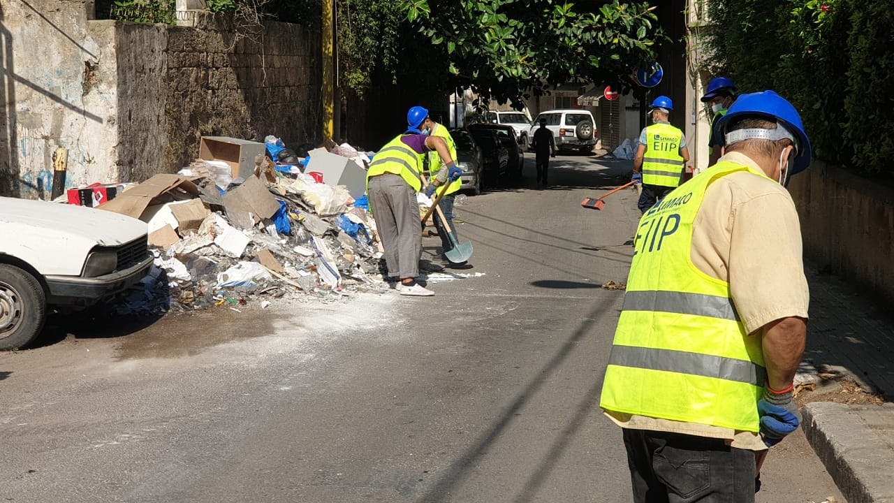 Workers clear rubble on Beirut street and wear blue hard hats and fluorescent jerseys with EIIP written on them.