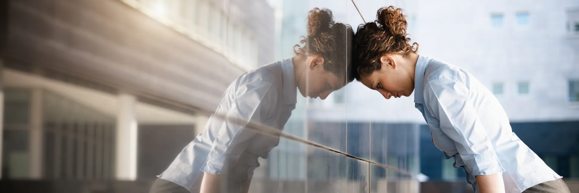 Adult woman leaning her head against a reflective wall outside an office building.