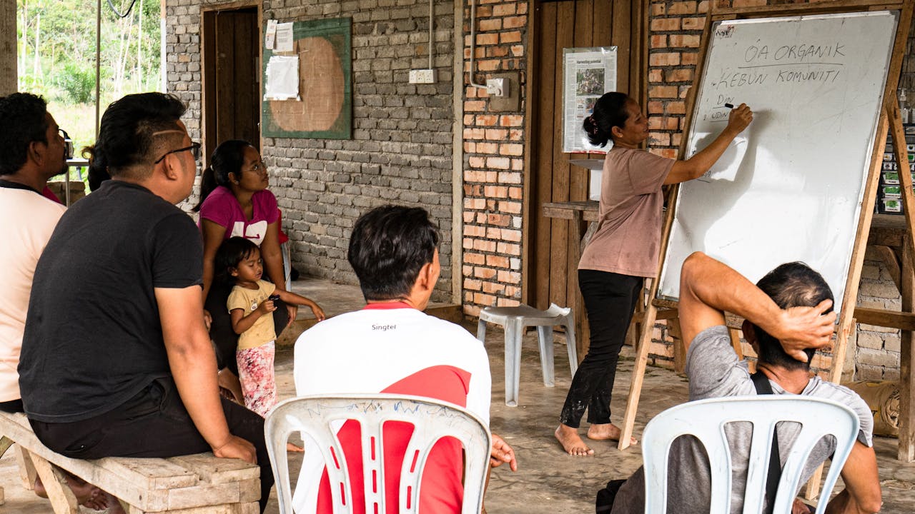 Maimun shares her knowledge with others in a community setting.  People are seated on a bench and chairs listening to her. She writes on a white board.