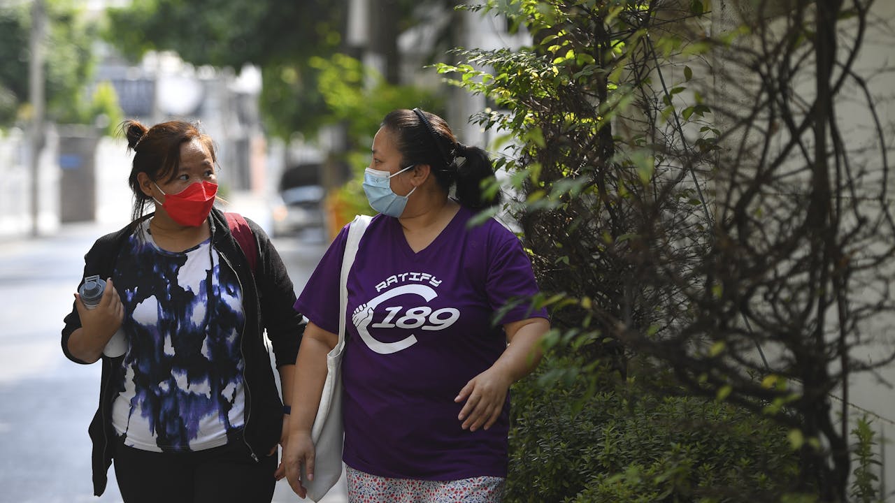Champa walks along a city sidewalk and talks with another migrant domestic worker.  They both wear masks. 