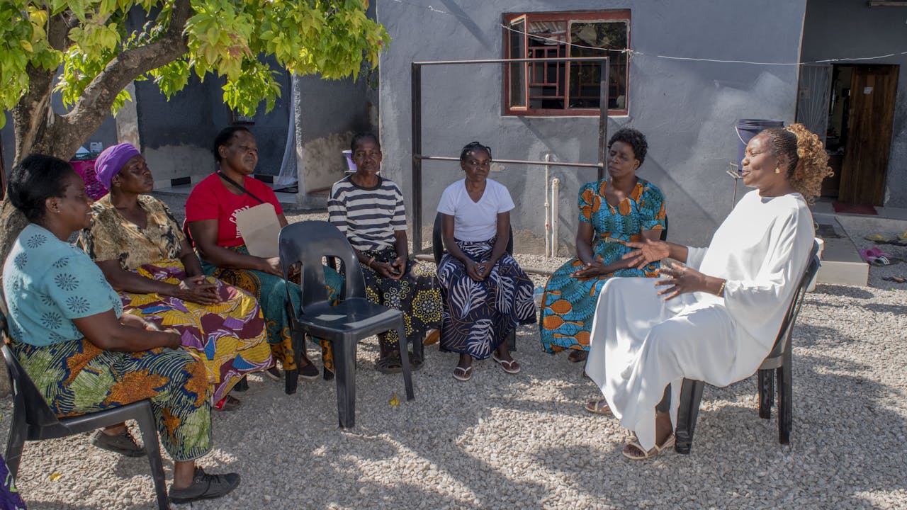 Siete personas sentadas fuera en círculo. Regina Katungu Chileshe está vestida de blanco y dirige una reunión de su grupo de apoyo comunitario frente al VIH. (2024)