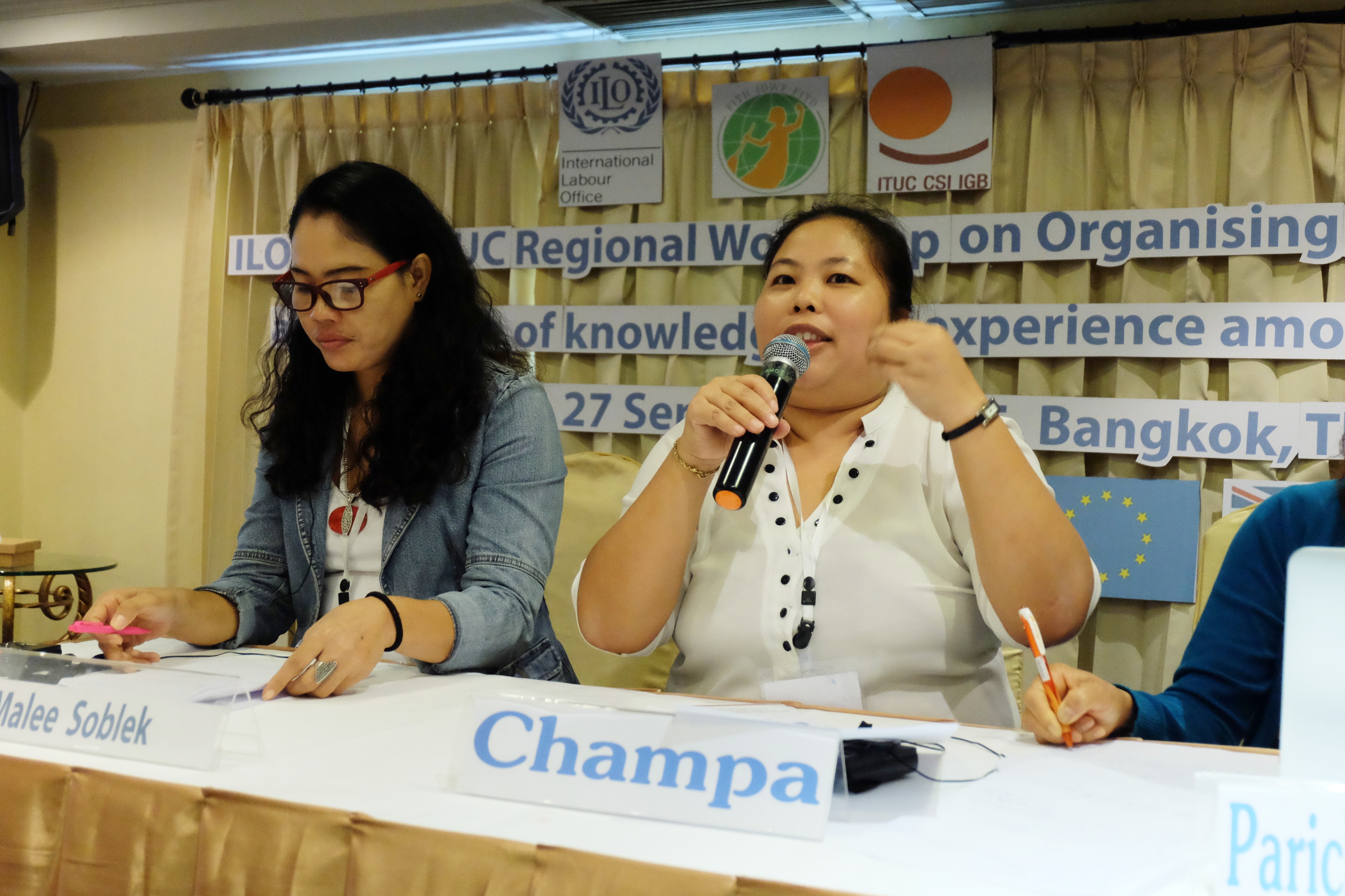 Champa est assise. Elle tient un micro et parle lors d'une réunion. Son nom figure sur une carte posée sur la table devant elle.