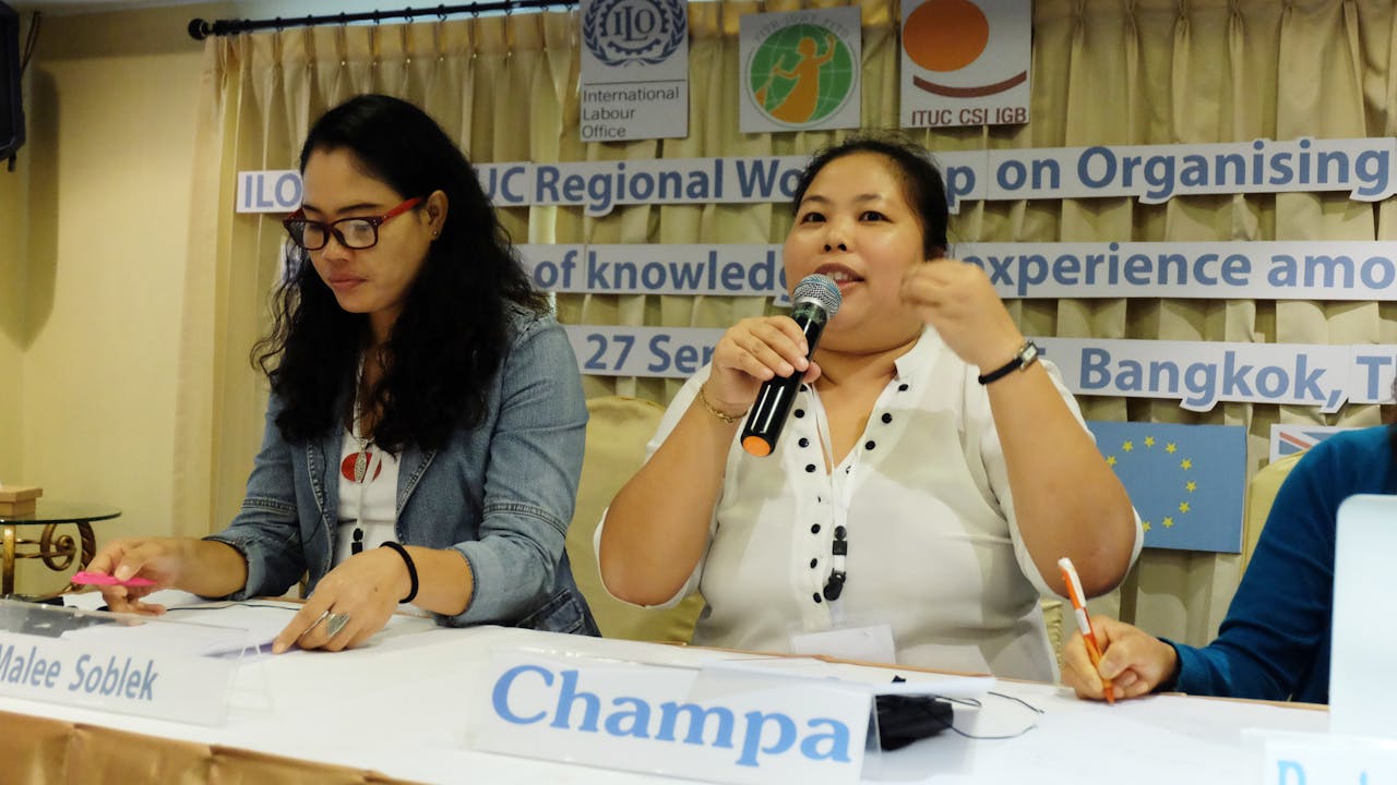 Champa is seated. She holds a microphone and speaks at a meeting. Her name is on a card on the table in front of her. 