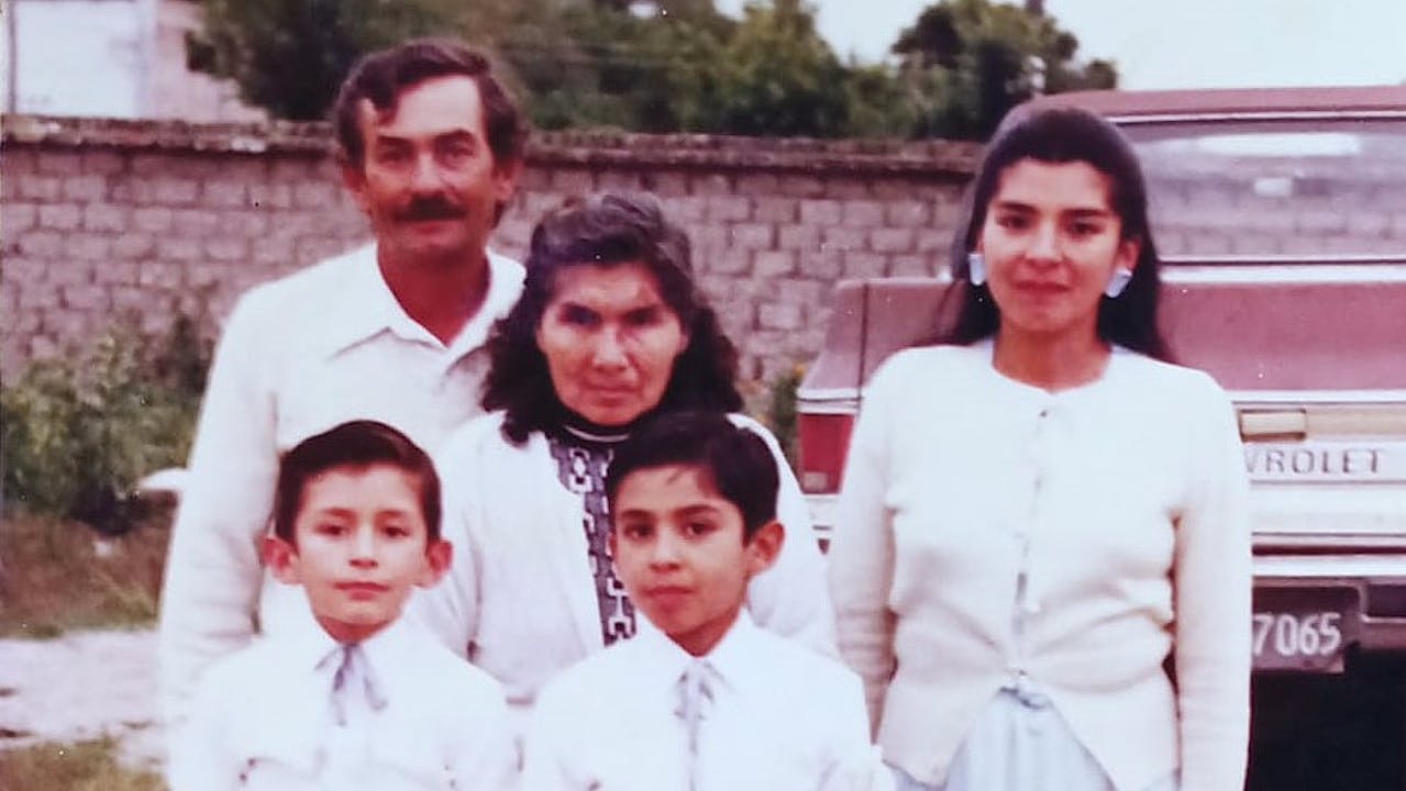 Daniel and his brother stand with their parents and grandmother outside. They all wear white. In the background is a brick wall and a Chevrolet pick-up truck. 