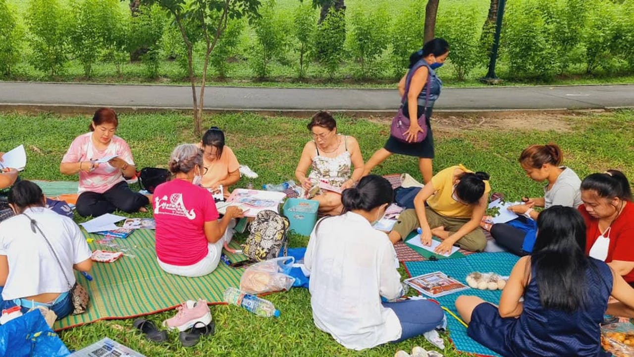 A group of women in a park sit and cut out pieces of paper and write on them. 