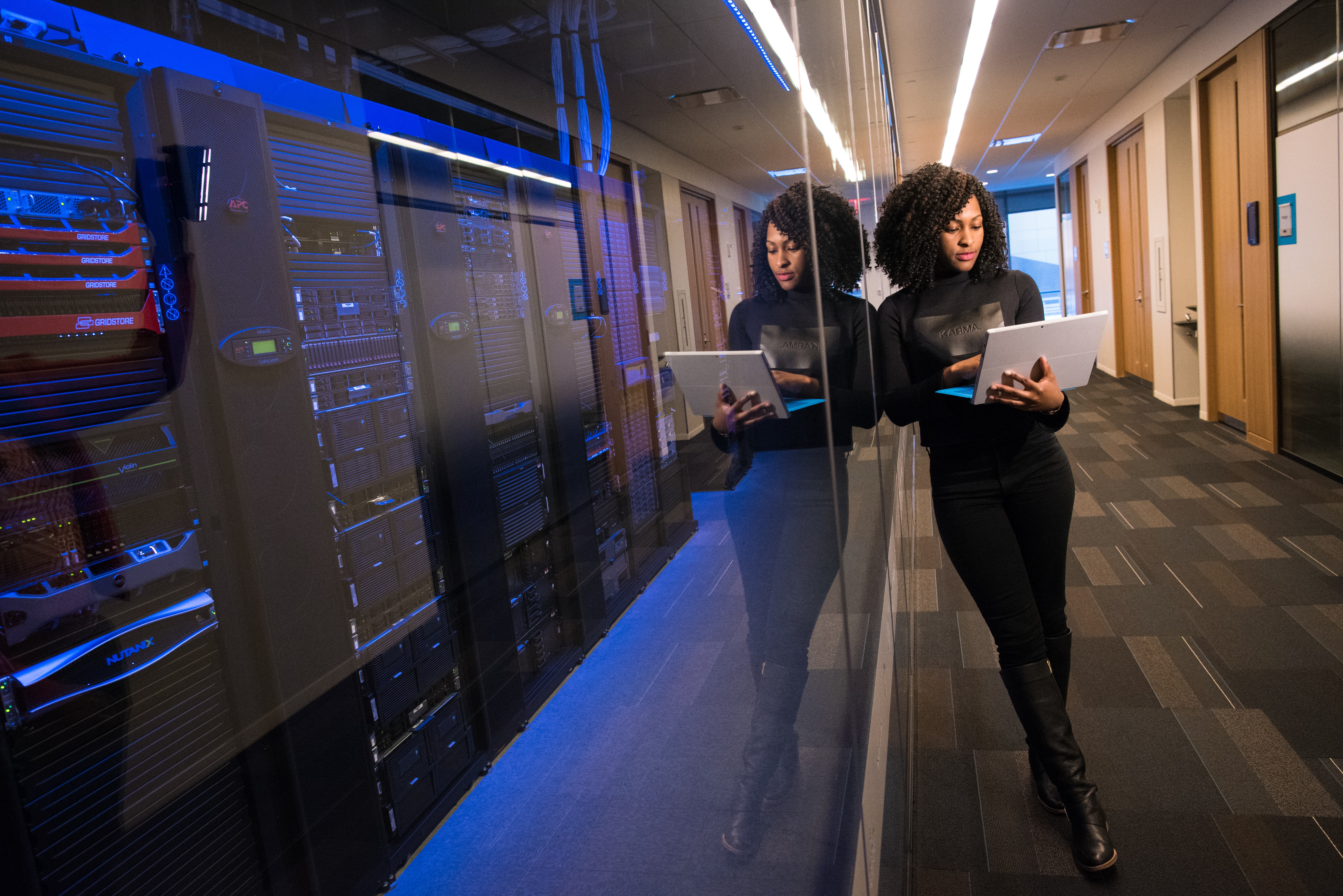 A woman holds a laptop which she is looking at. She is in an office hallway and leans on a transparent wall, behind which are a row of computer servers.