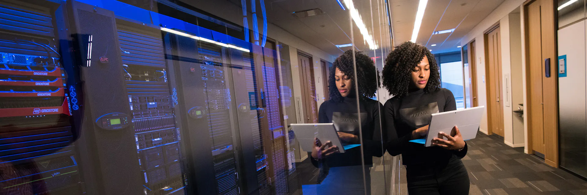 A woman holds a laptop which she is looking at. She is in an office hallway and leans on a transparent wall, behind which are a row of computer servers.