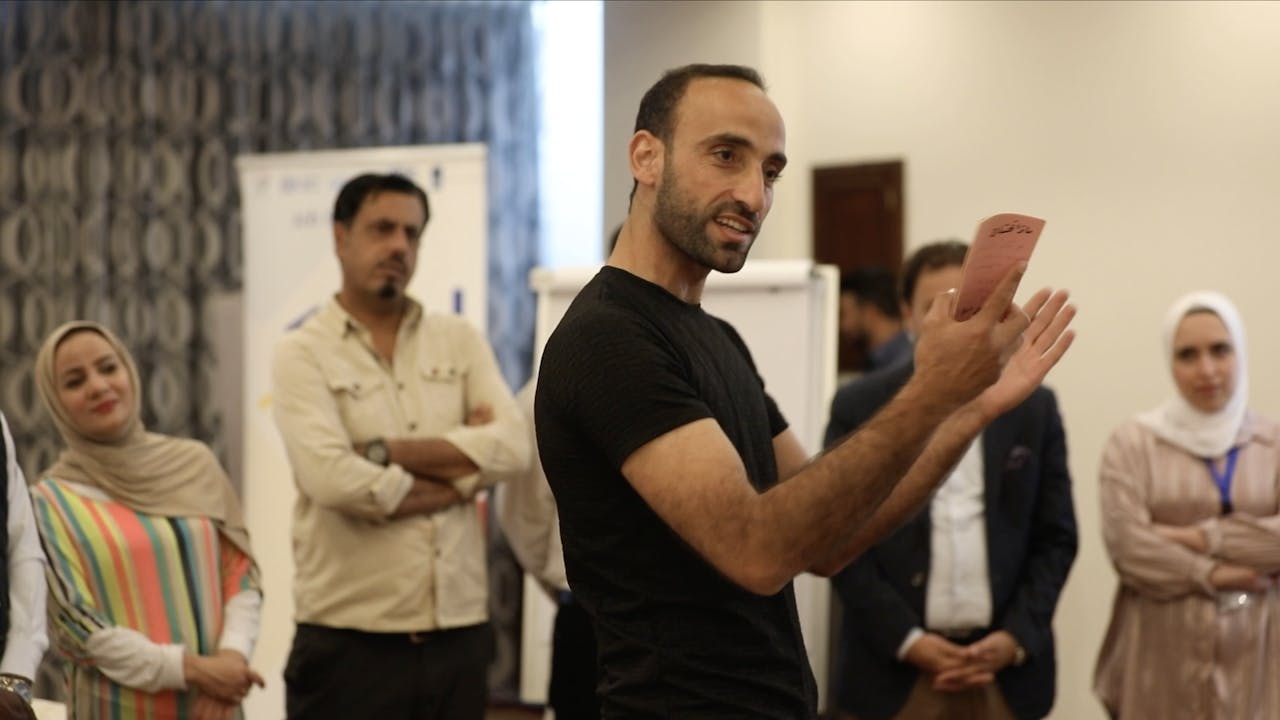 Abdel Halim al Qasir stands in the middle of a group of people in a room, he holds up a document and is explaining something.  
