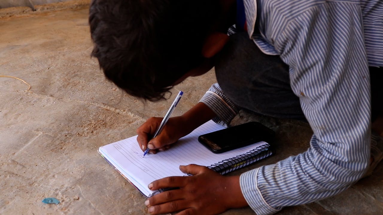 Fatima’s son is crouched over a mobile phone and writes in his school notebook.