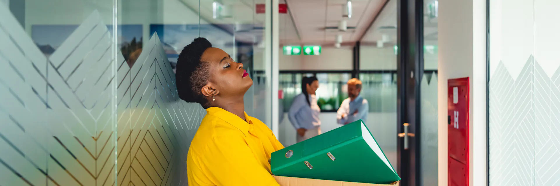 A young woman stands in a corridor holding a box of office supplies. She leans her head back on the office hallway wall. Her eyes are closed and she looks unhappy.