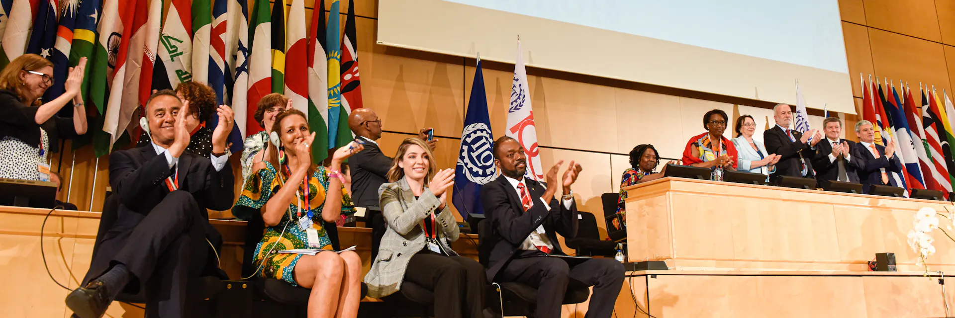 Delegates smile and clap after the adoption of Convention 190 on the elimination of violence and harassment in the world of work. A screen behind them shows the results of the adoption vote. The adoption took place in the UN General Assembly Hall at the 108th International Labour Conference on June 21, 2019.