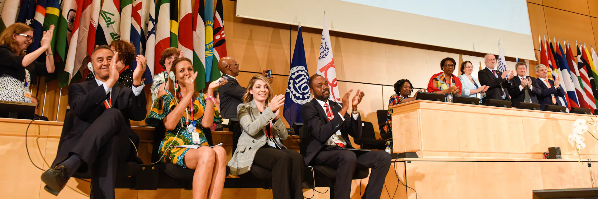 Delegates smile and clap after the adoption of Convention 190 on the elimination of violence and harassment in the world of work. A screen behind them shows the results of the adoption vote. The adoption took place in the UN General Assembly Hall at the 108th International Labour Conference on June 21, 2019.
