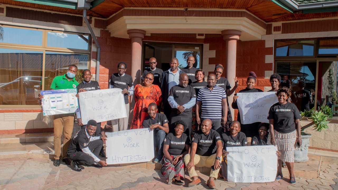 Regina Katungu Chileshe, sentada en la parte inferior derecha, sostiene un cartel que reza "ILO VCT@WORK HIV testing", que aboga por el asesoramiento y la realización de pruebas de forma voluntaria en el trabajo. Posa para la cámara en el exterior de un edificio con otros participantes en el programa de formación de la OIT. (2024)
