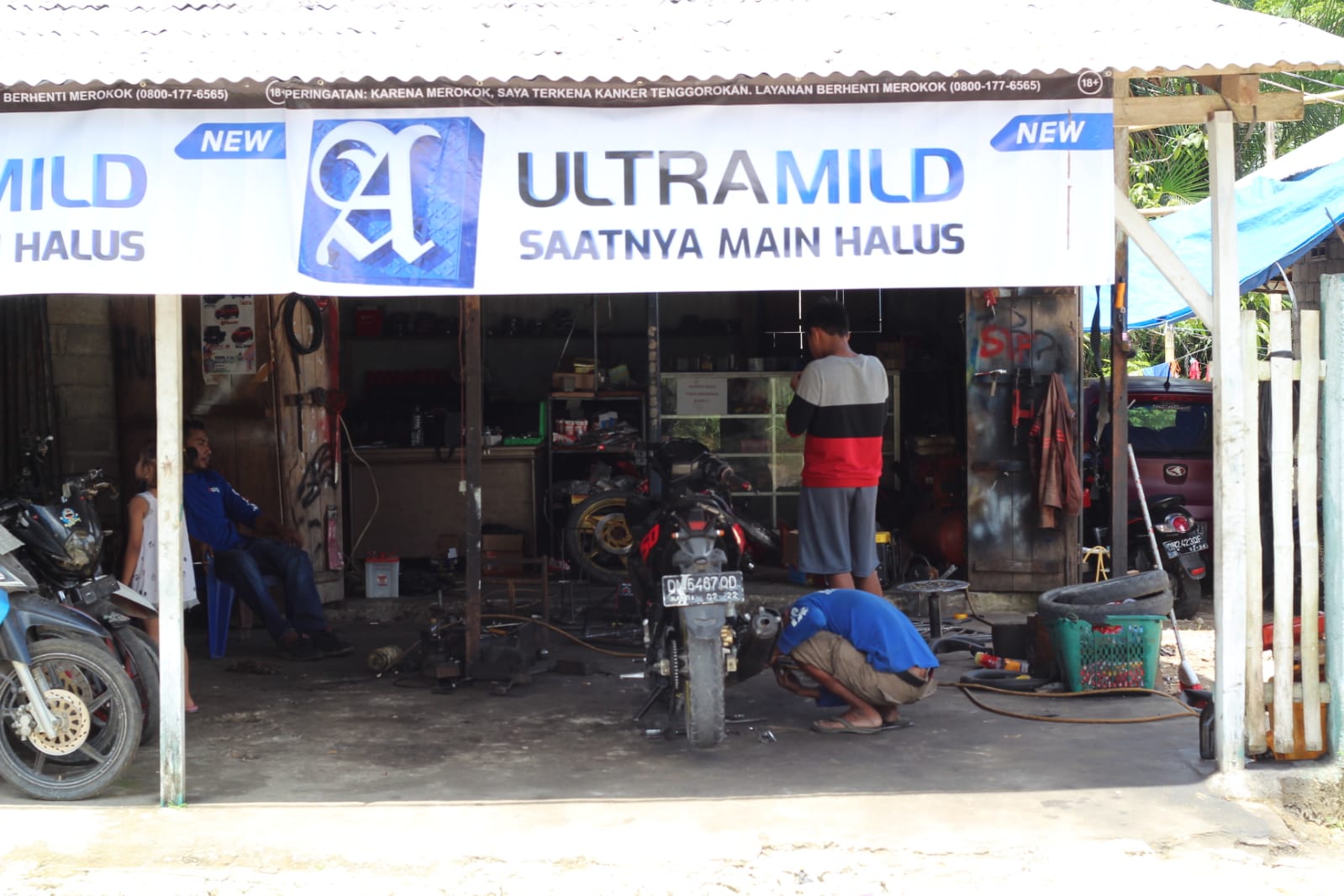 Extérieur de l’atelier de réparation de motos et de moteurs de bateaux. Un mécanicien est accroupi au sol et travaille sur une moto. Juanda est assis sur une chaise et l’observe.