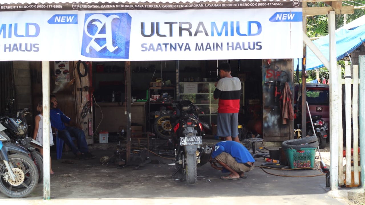 The exterior of the motorbike and boat engine repair shop. A mechanic is crouched on the ground and works on a motorbike. Juanda sits on a chair and looks on.