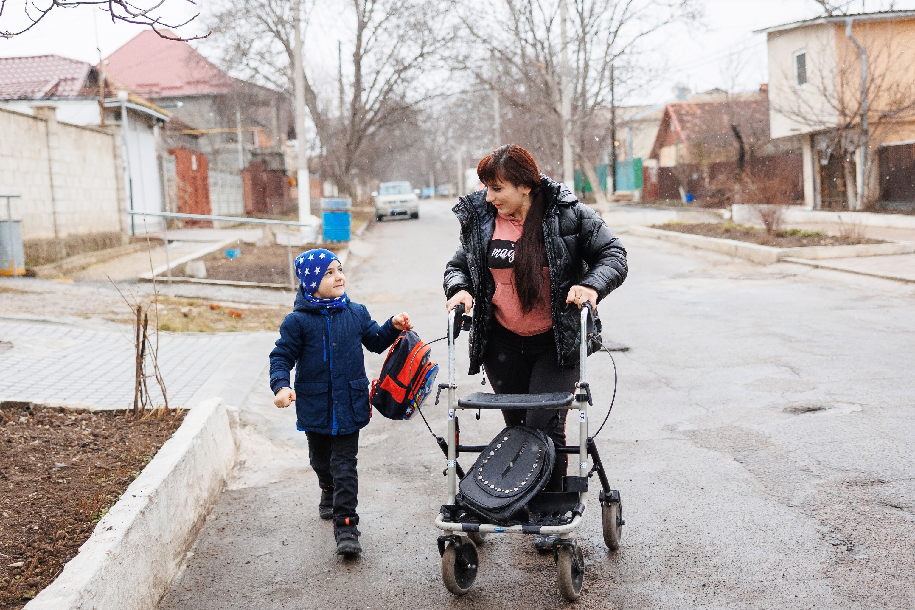 Elena and her son Maxim walk along a street on a grey, cold day. They look at each other with tenderness. Elena uses a walker. Maxim holds a backpack. 