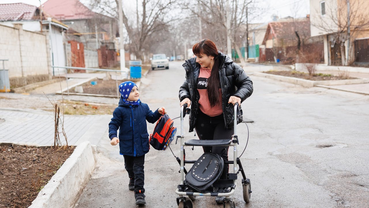 Elena and her son Maxim walk along a street on a grey, cold day. They look at each other with tenderness. Elena uses a walker. Maxim holds a backpack. 