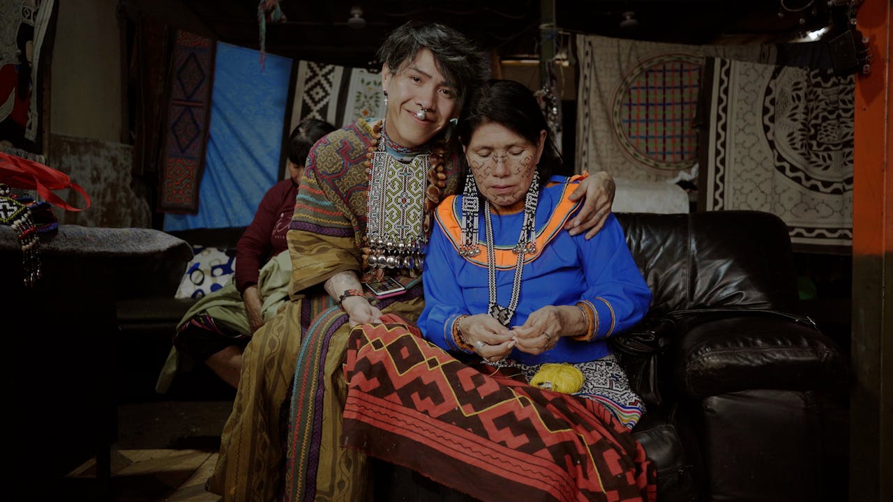 Olinda and Roné sit side by side. Olinda wears glasses and is working on a piece of fabric. Roné has his arm around his mother and smiles to the camera.