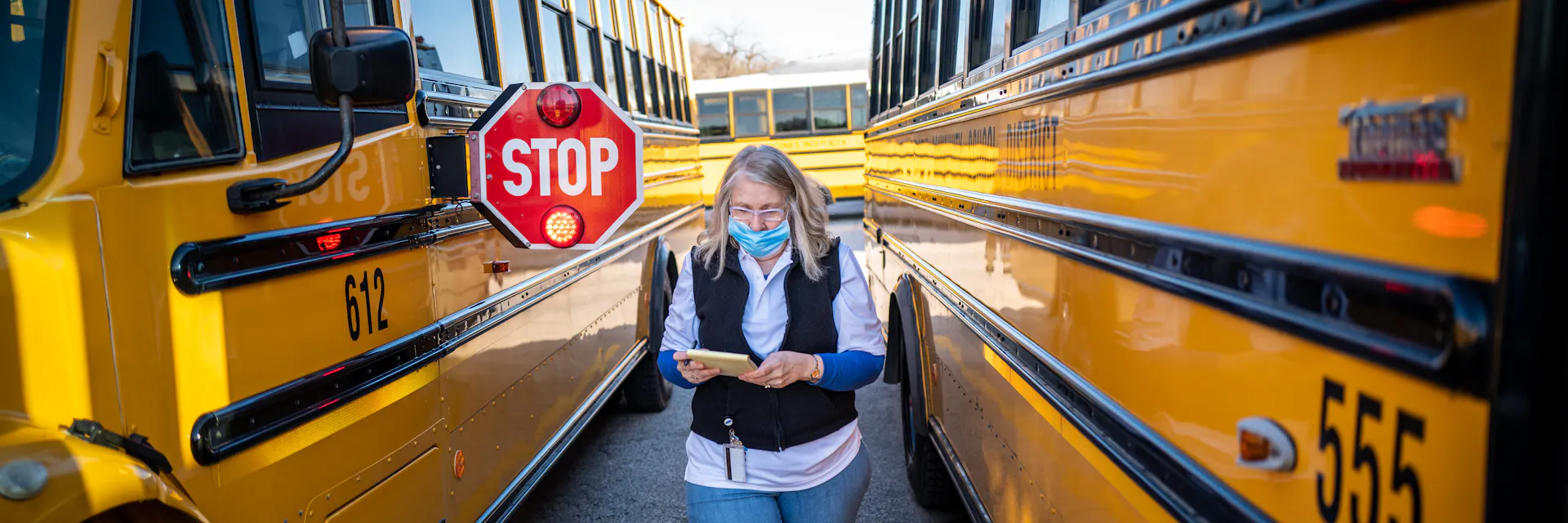 A female teacher in the United States during the COVID pandemic wears a mask and holds a notepad. She stands between two orange school buses.