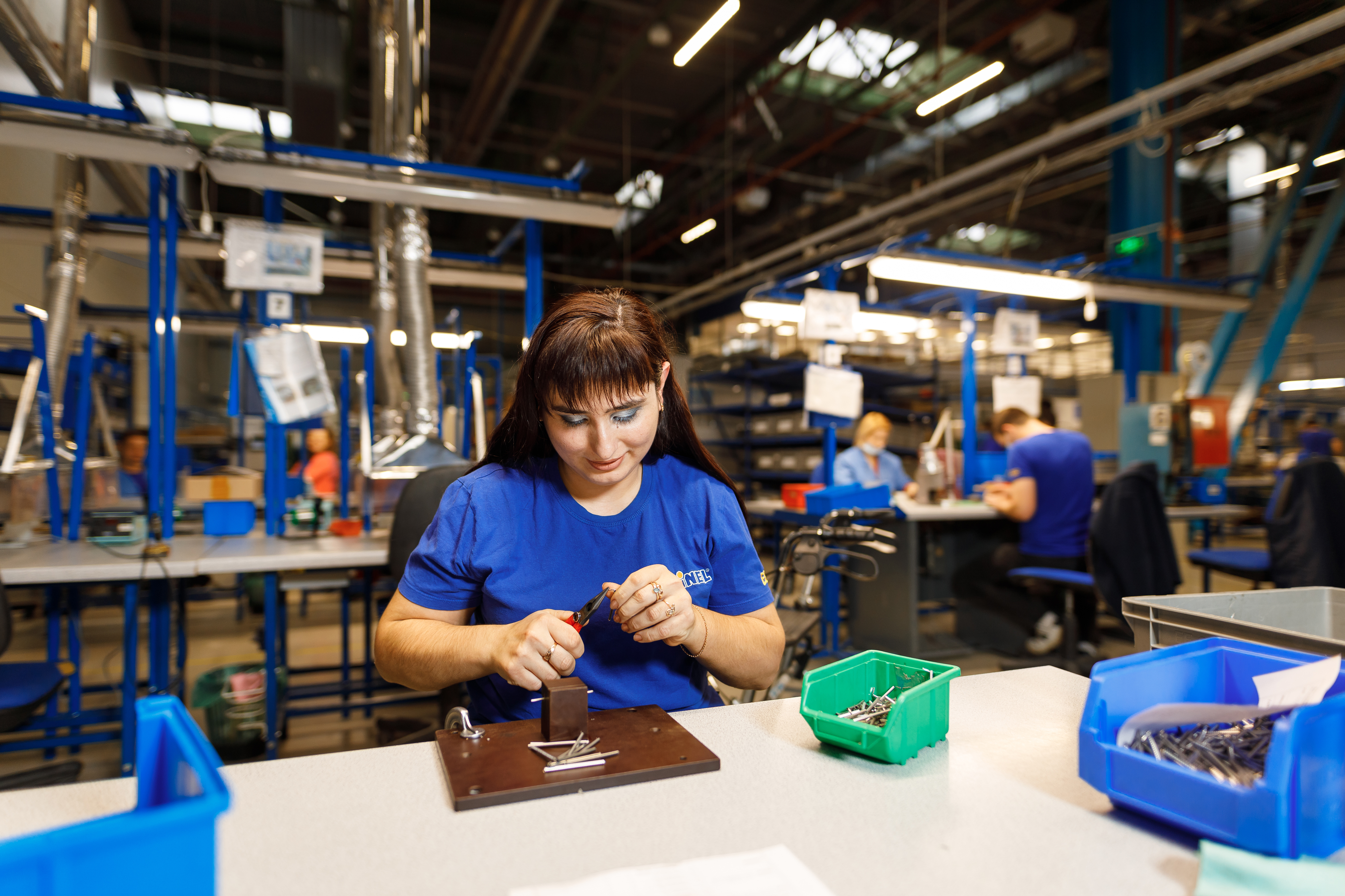 Elena sits at a large table assembling parts. She is in a workshop with high ceilings. Other workers are in the background at another work station.
