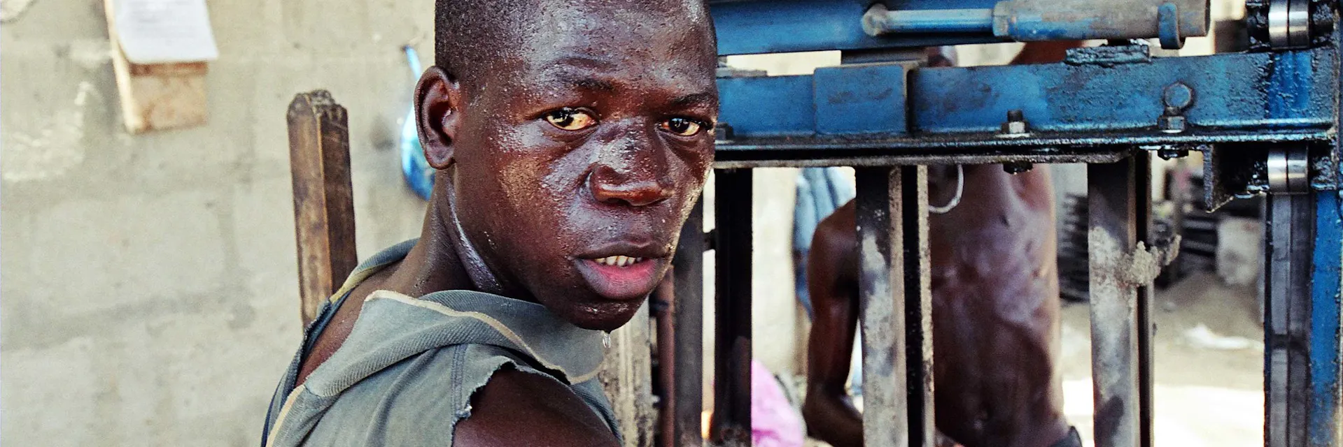 Un ouvrier est assis à côté de sa machine de travail dans une usine de briques de ciment en Tanzanie. Il regarde la caméra. Il a visiblement très chaud. Date: 2003-10