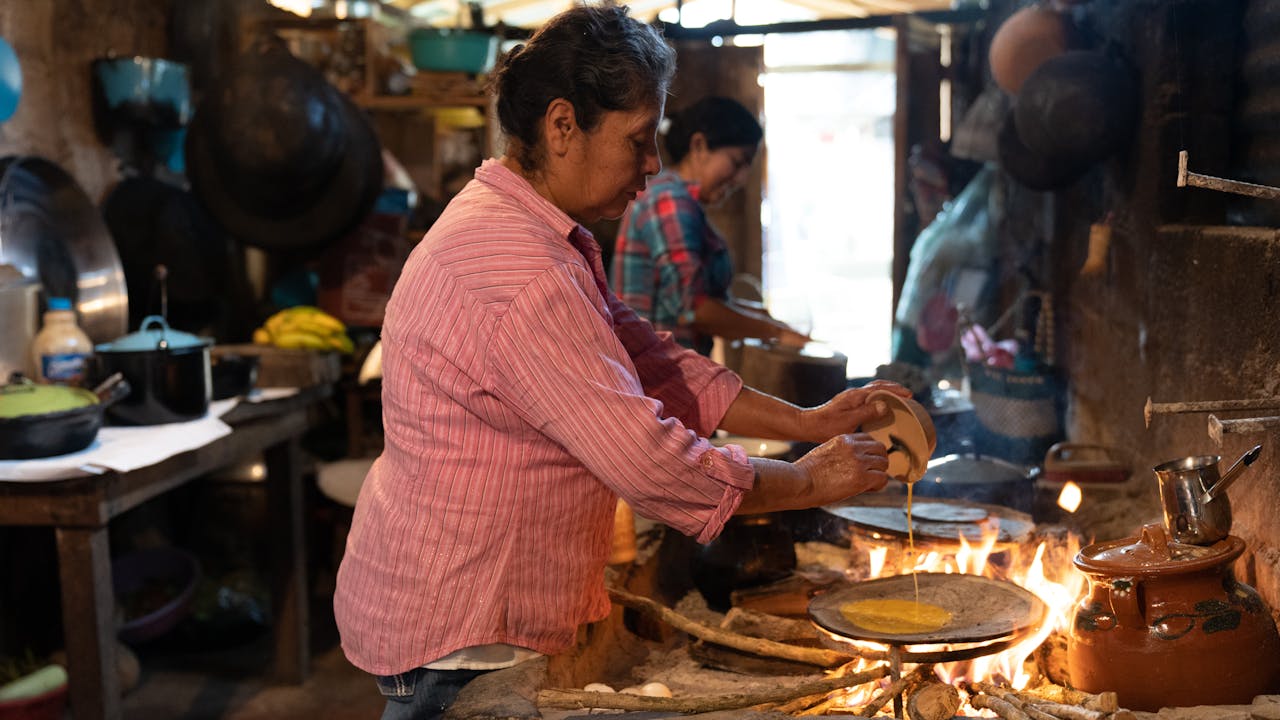Briseida presses a tortilla into shape in the background. In the foreground an older woman pours whisked egg onto a tortilla, that is cooking over an open fire.