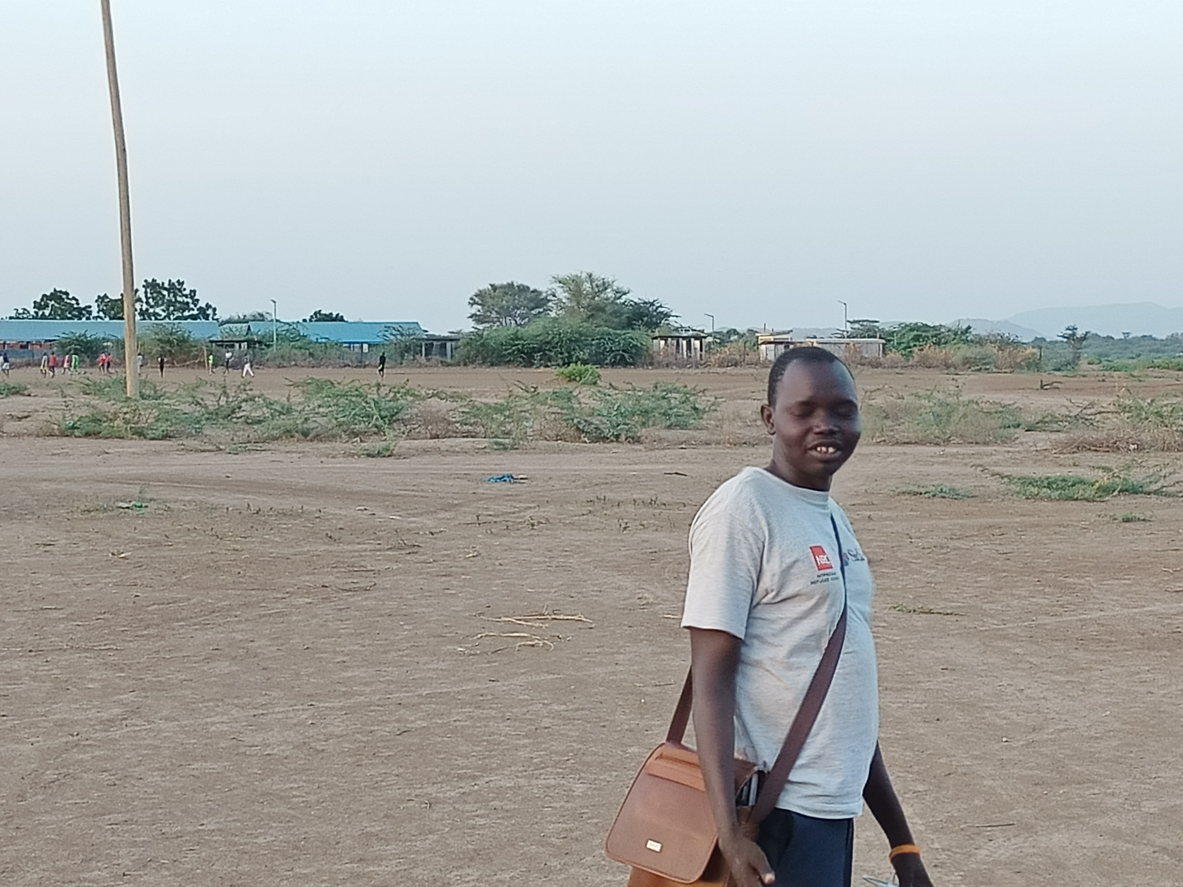 Thon de pie, con un árido paisaje con poca vegetación y edificios del campamento de Kakuma al fondo.