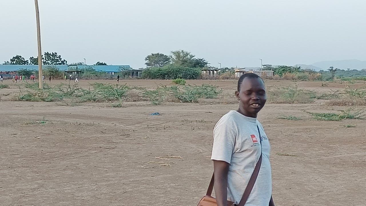 Thon Mabior Jok stands in a dry landscape with little vegetation, with buildings of Kakuma camp in the background.