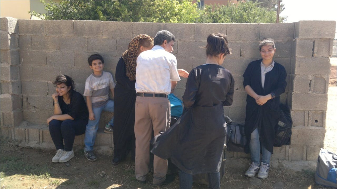 Jin Dawod’s family stand outside next to a wall of concrete blocks with their bags.  Three children, including Jin, look at the camera.  The adults – two women and a man - have their back to the camera and are deep in discussion.