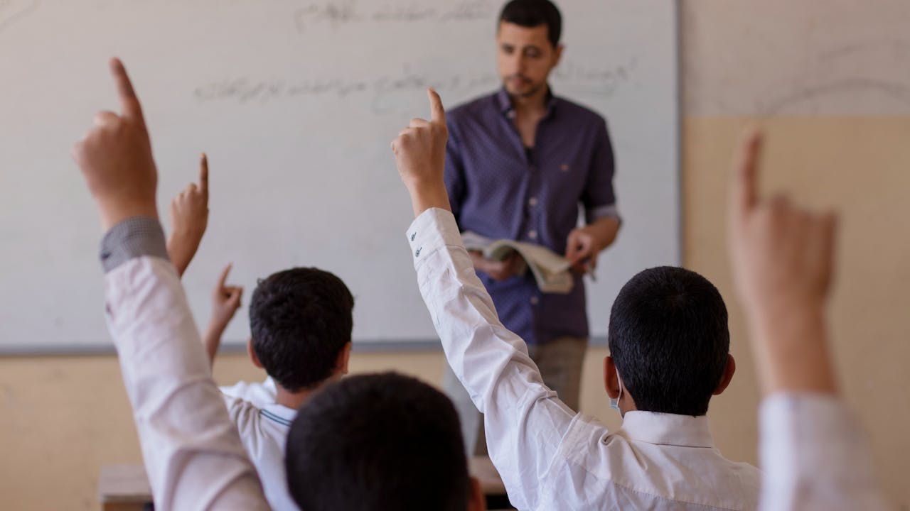 Des enfants, dont Akram, lèvent la main dans une salle de classe face à leur enseignant qui se tient devant un tableau blanc.