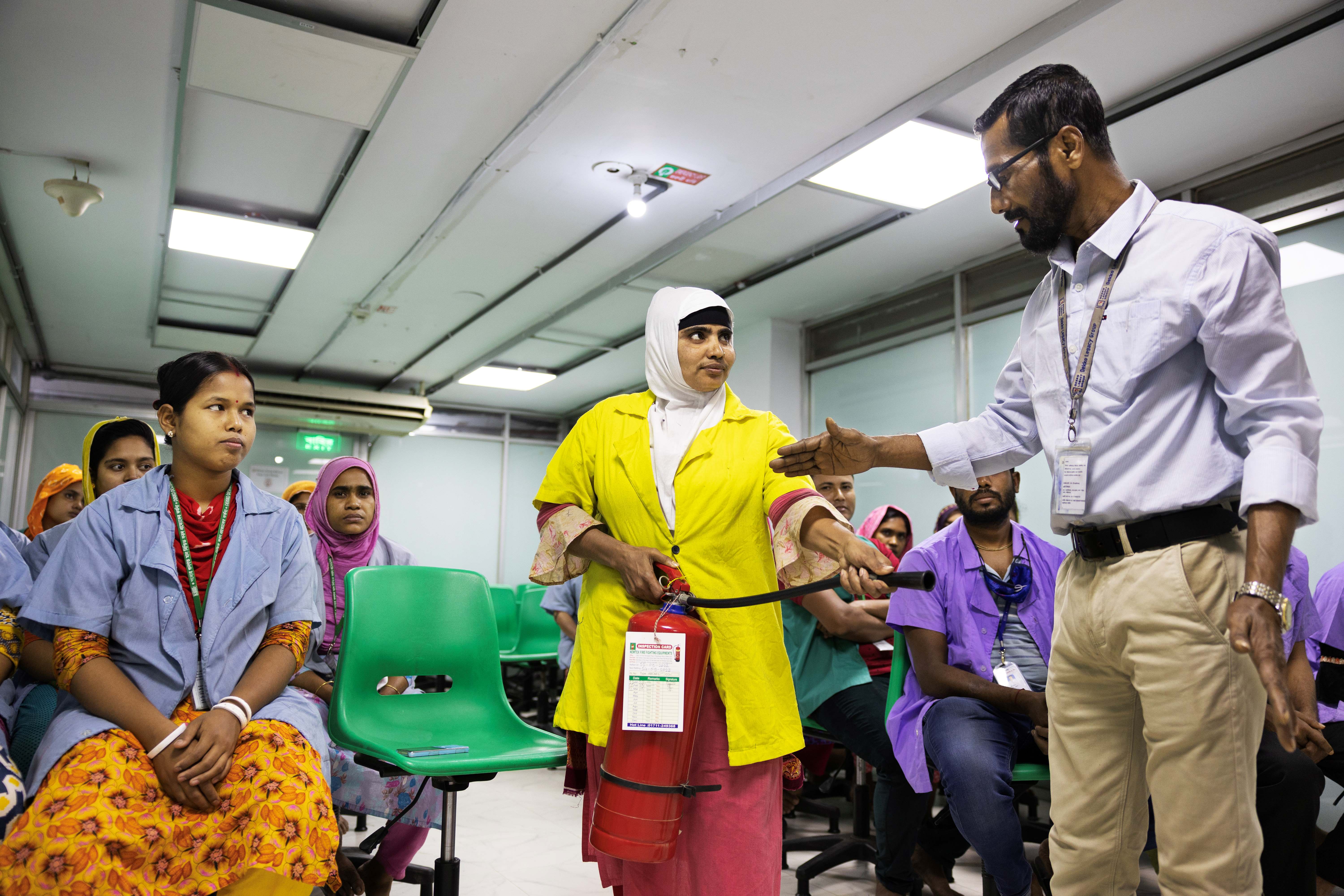 Mohamad Abdur Rob shows  a woman how to use a fire extinguisher.  Other workers in the training workshop are seated and look on. 