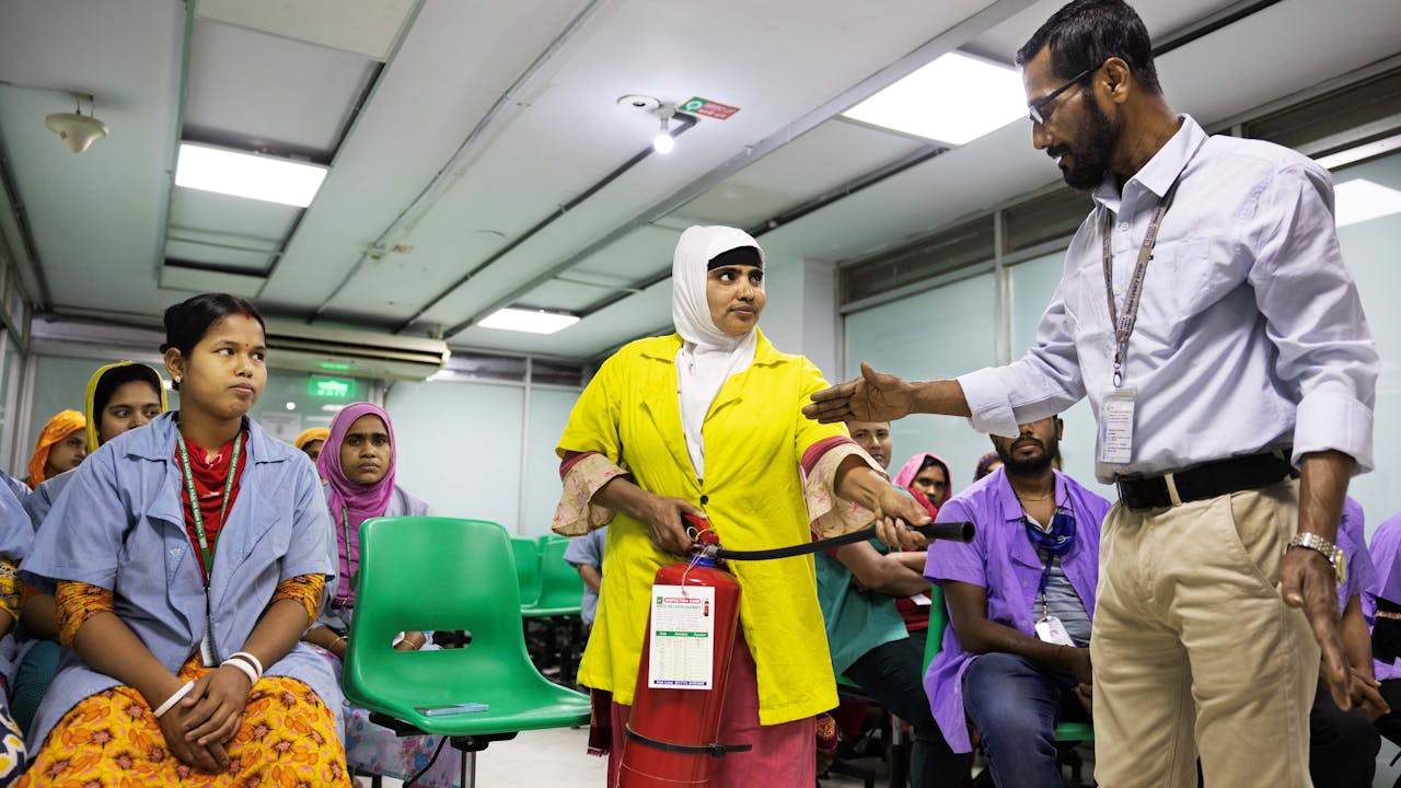 Mohamad Abdur Rob shows  a woman how to use a fire extinguisher.  Other workers in the training workshop are seated and look on. 