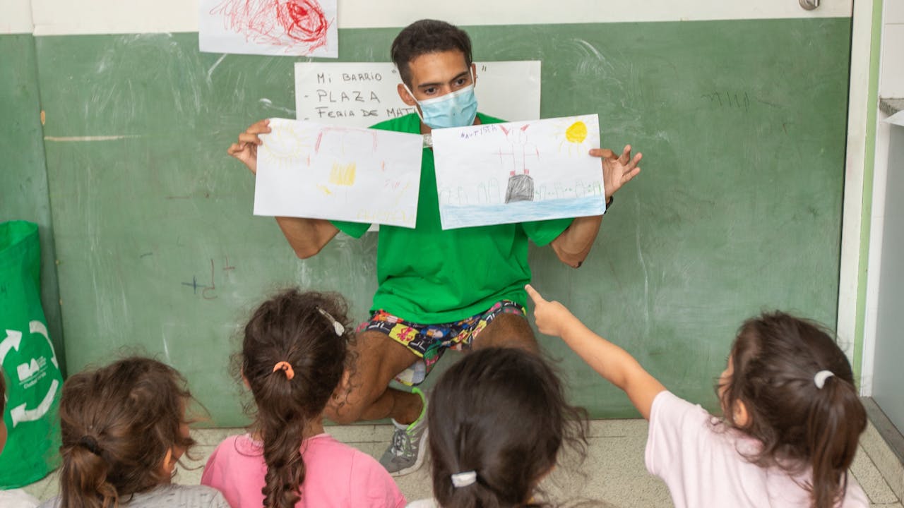 In a classroom Federico Trotta holds two children’s paintings in his hands as the children sit in a row and interact with him.