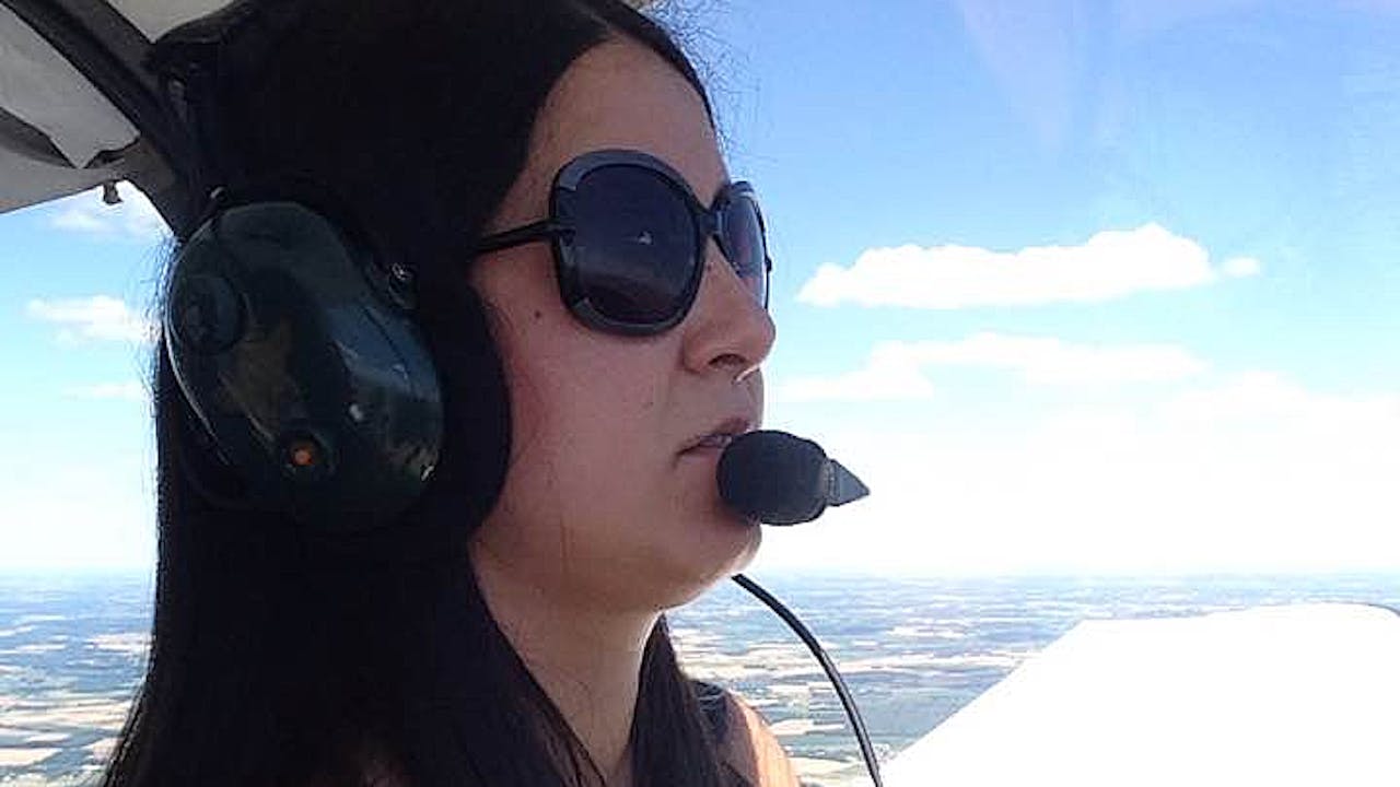 A close-up of Güler Koca wearing a headset with built-in microphone and sunglasses in the cockpit of a small plane. She is piloting the plane. In the background we see the blue sky and landscape far below stretching out into the distance.