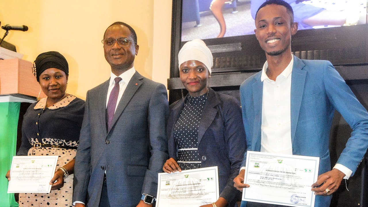 Cissé Mabré and two other training participants hold up their SIYB training certificates. They are smartly dressed and stand next to a man in a suit.