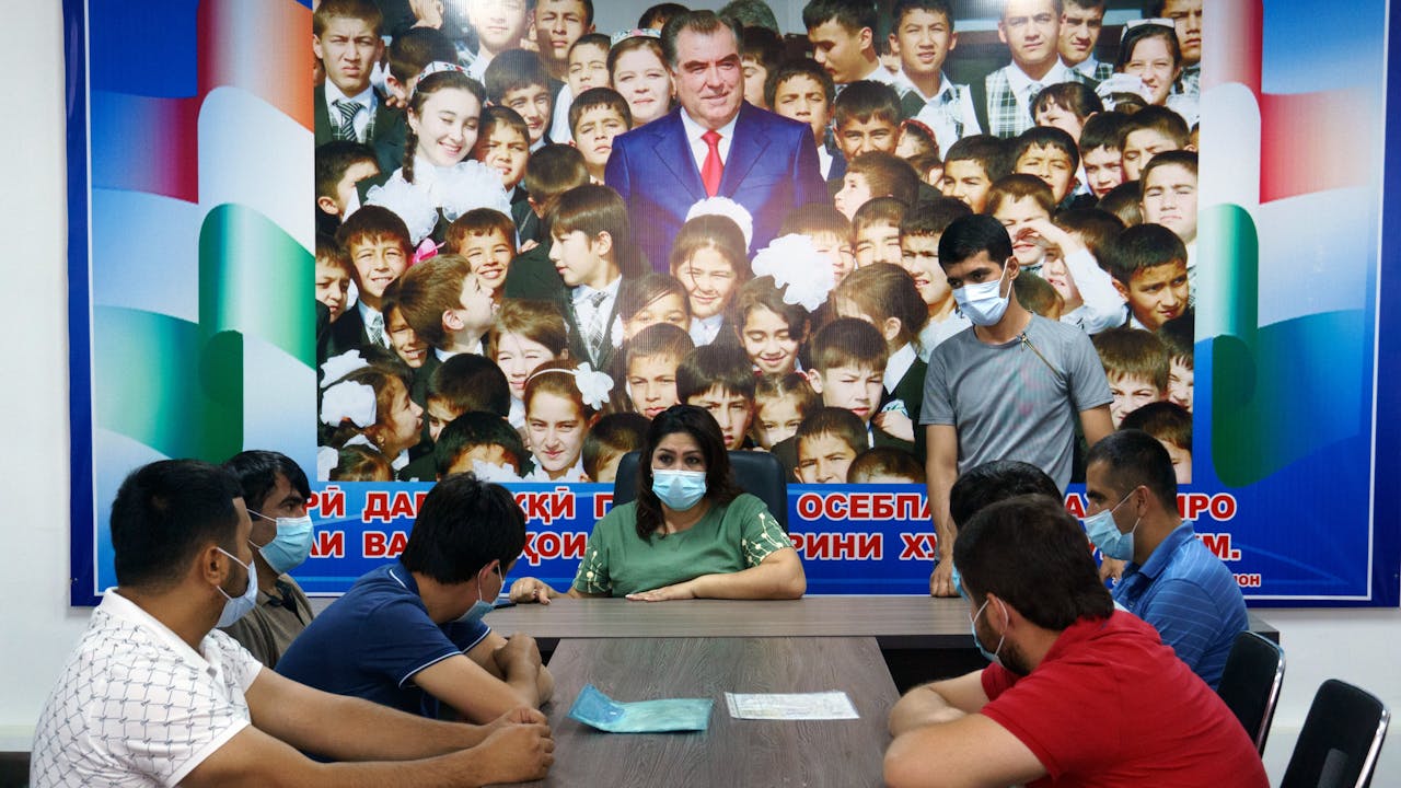 Takhmina sits at the head of a meeting table with masked taxi drivers, all men, looking on. Behind them is a giant poster of Tajikistan’s President, Emomali Rahmon, standing in a crowd of children.