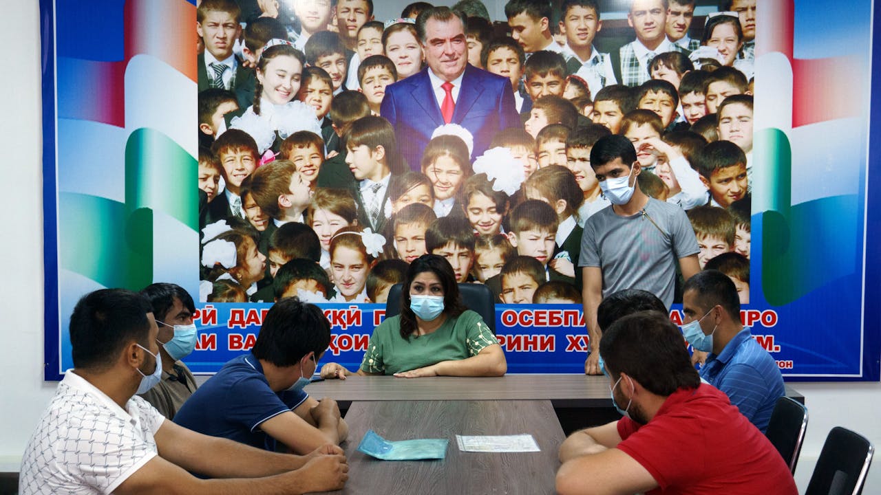 Takhmina preside una mesa de reunión con taxistas que llevan mascarilla, todos hombres, mientras la observan. Tras ellos hay un cuadro de gran tamaño del presidente de Tayikistán, Emomali Rahmon, de pie entre una multitud de niños.