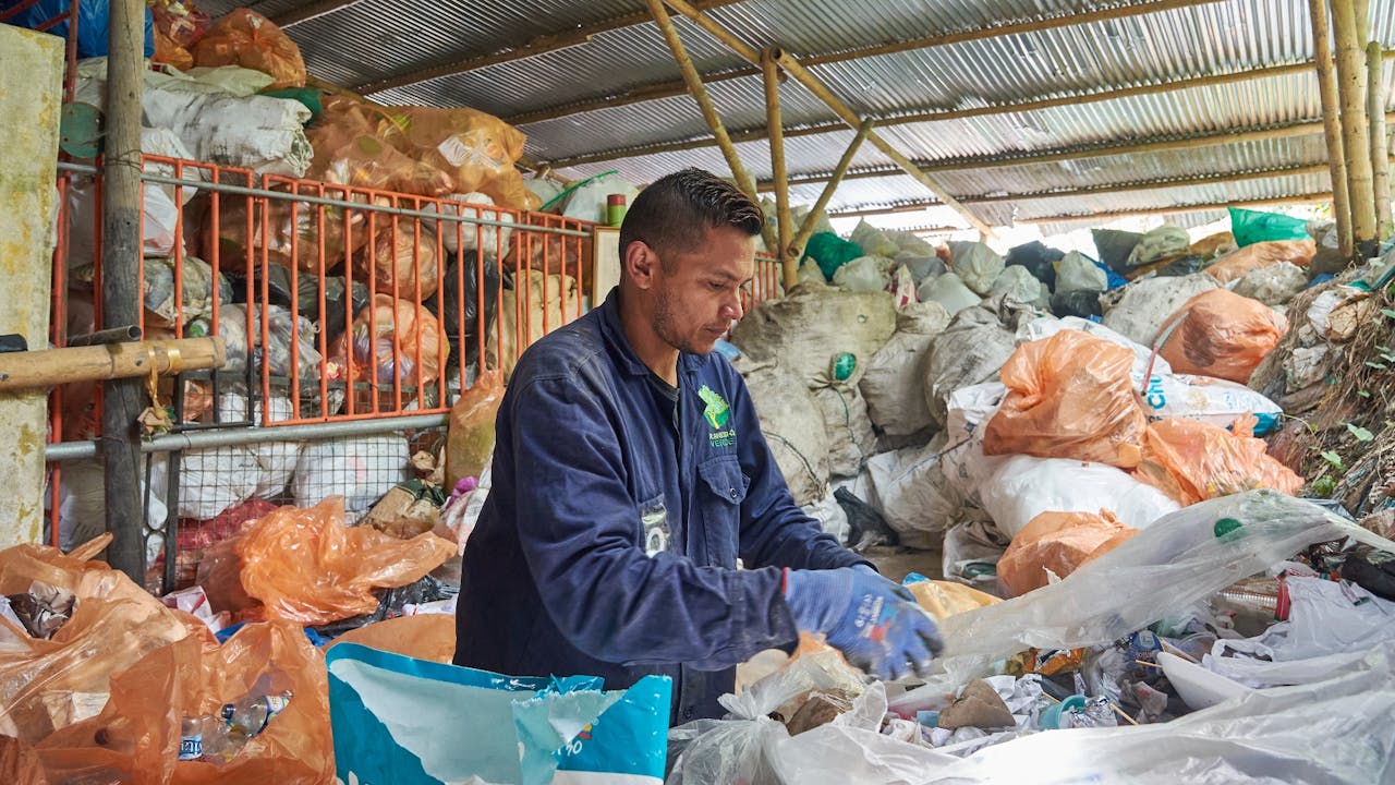 A waste picker sorts waste in the recycling warehouse. He wears blue gloves and blue overalls.