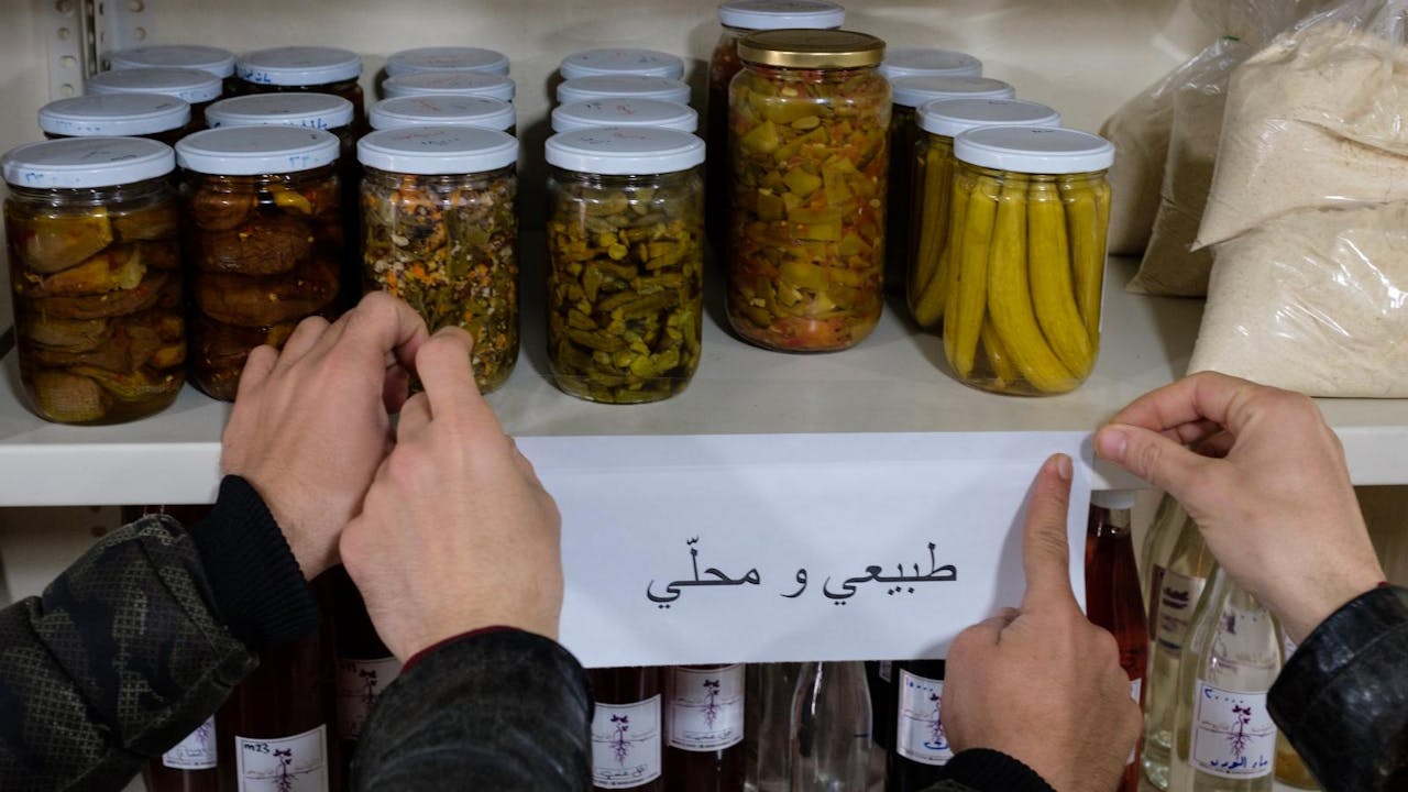 Dikkeneh Consumer Cooperative shopkeepers attach a sign in Arabic, which reads "natural and local", to a shelf lined with pickled food in glass jars.