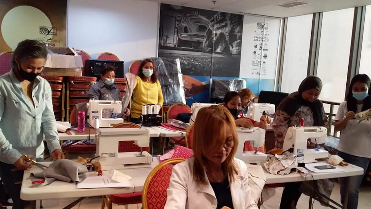 Wide shot of a room filled with tables where Filipino migrant domestic workers are busy sewing using sewing machines.