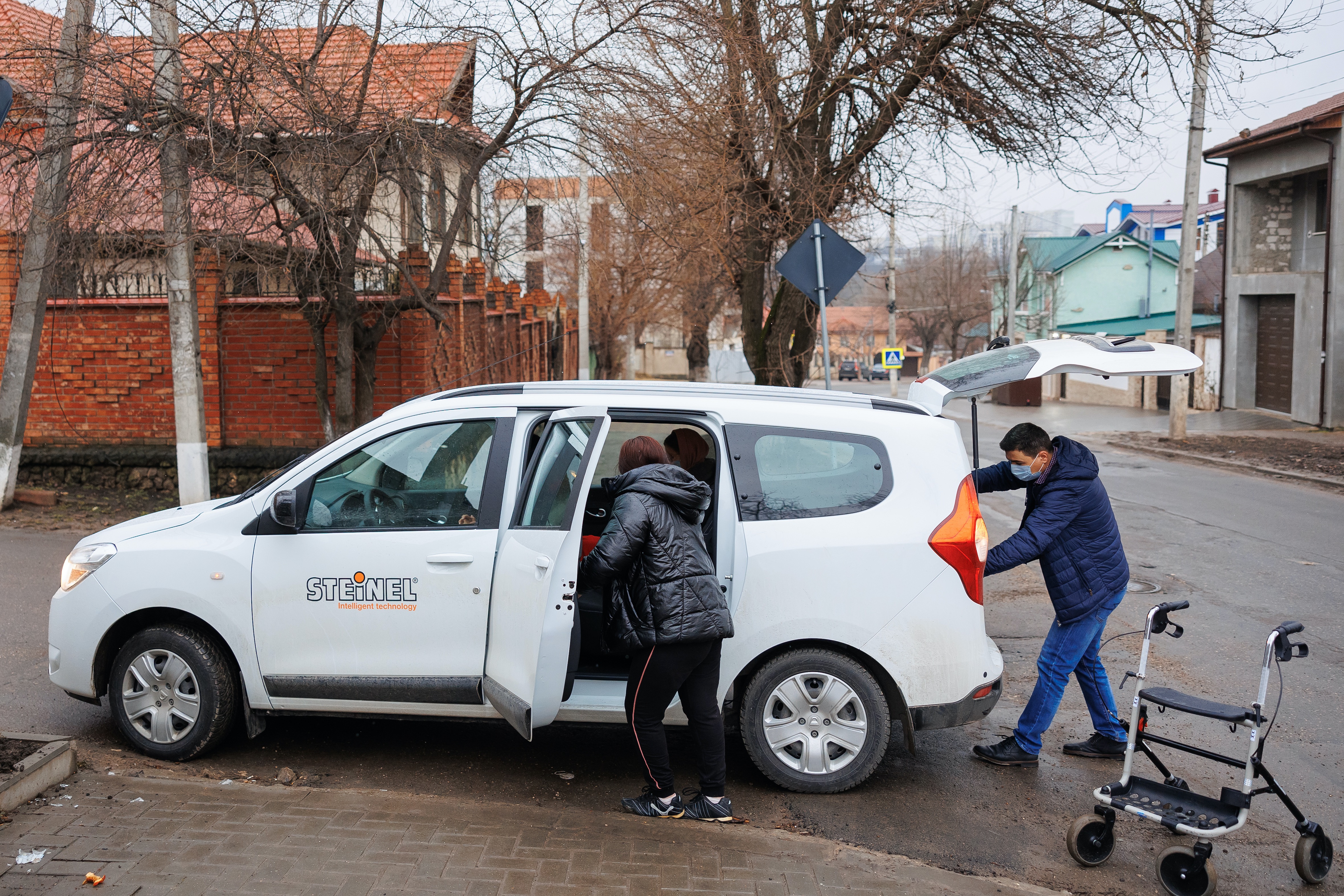 Elena gets into the back seat of a van belonging to her employer, while the driver prepares to put her walker in the trunk.