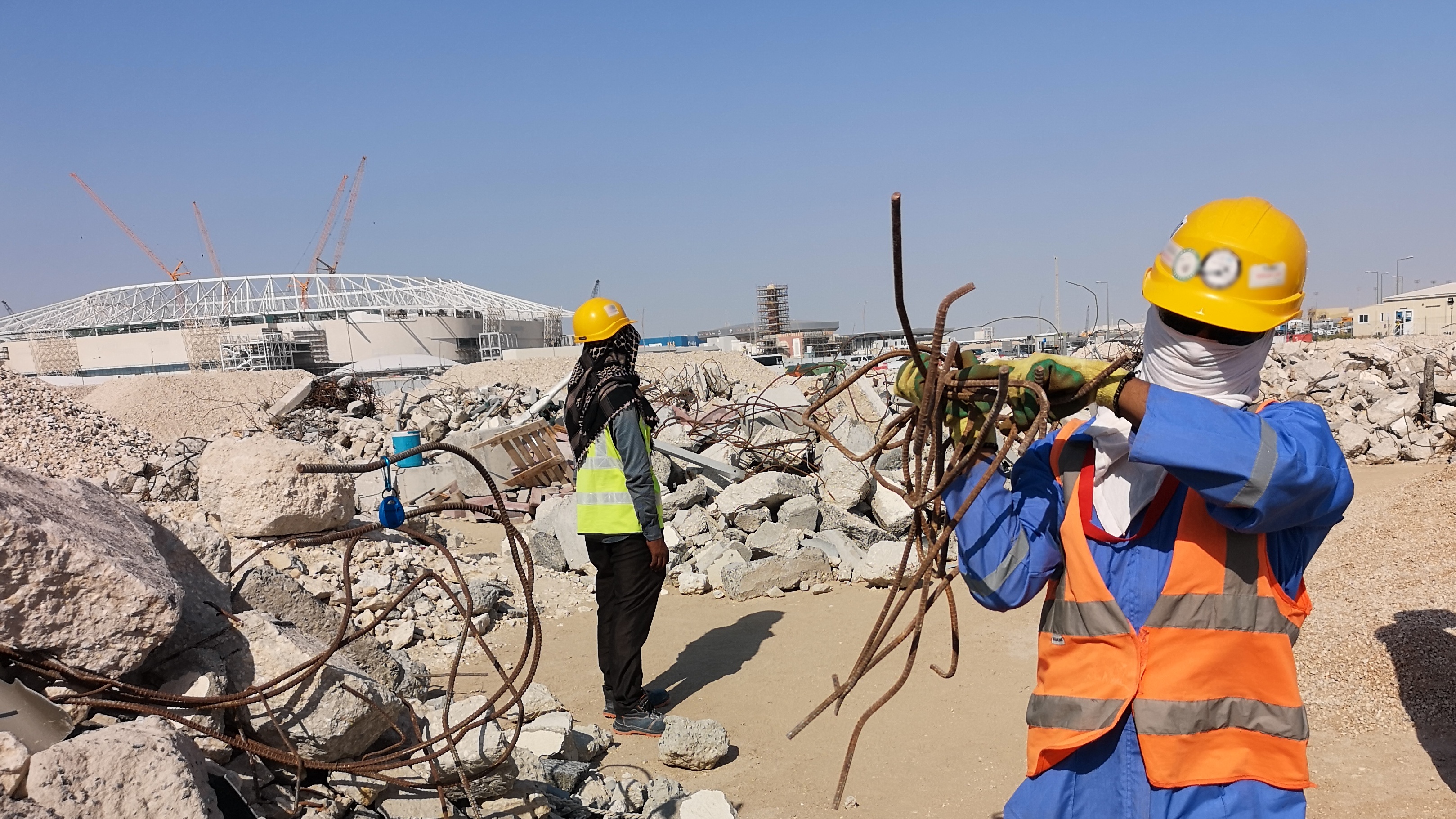 Construction worker wearing scarf around face and yellow hard hat carries pile of metal rods.  A stadium in mid-construction is in the background.