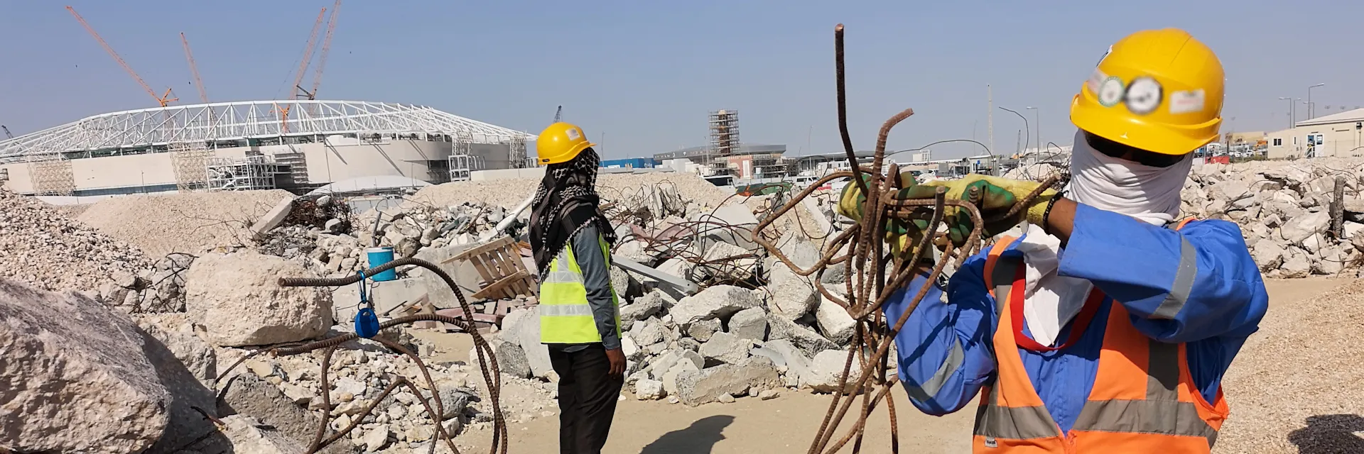 Construction worker wearing scarf around face and yellow hard hat carries pile of metal rods. A stadium in mid-construction is in the background.