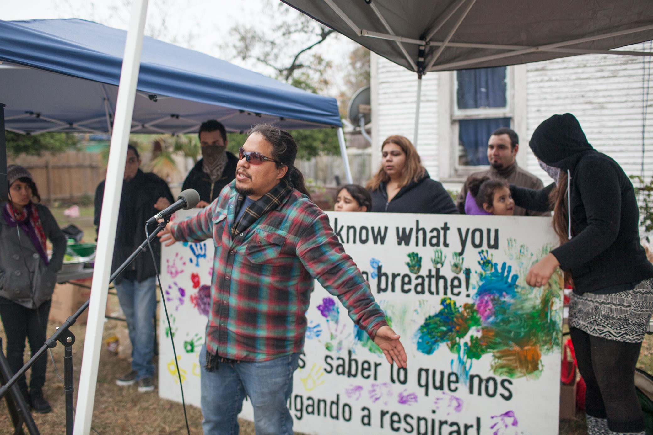 Bryan Parras speaks in front of a microphone at an outdoor event with a banner behind him where it’s written: "We demand to know what you are forcing us to breathe!"