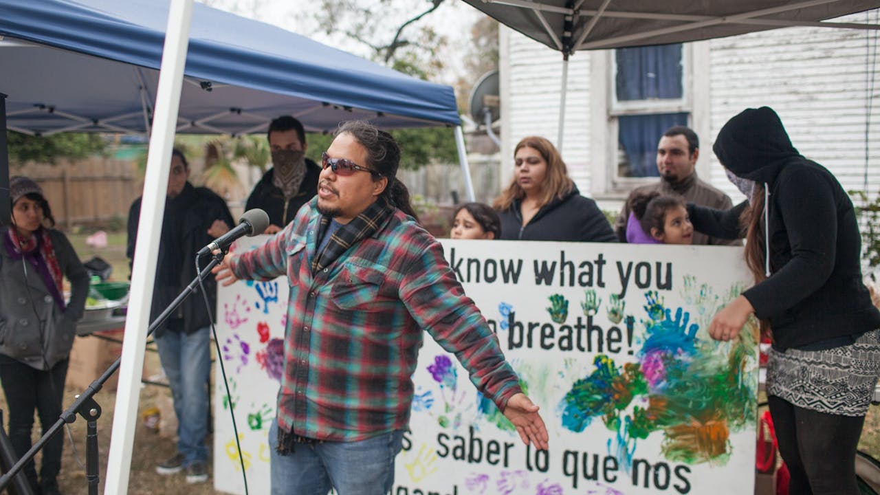 Bryan Parras speaks in front of a microphone at an outdoor event with a banner behind him where it’s written: "We demand to know what you are forcing us to breathe!"