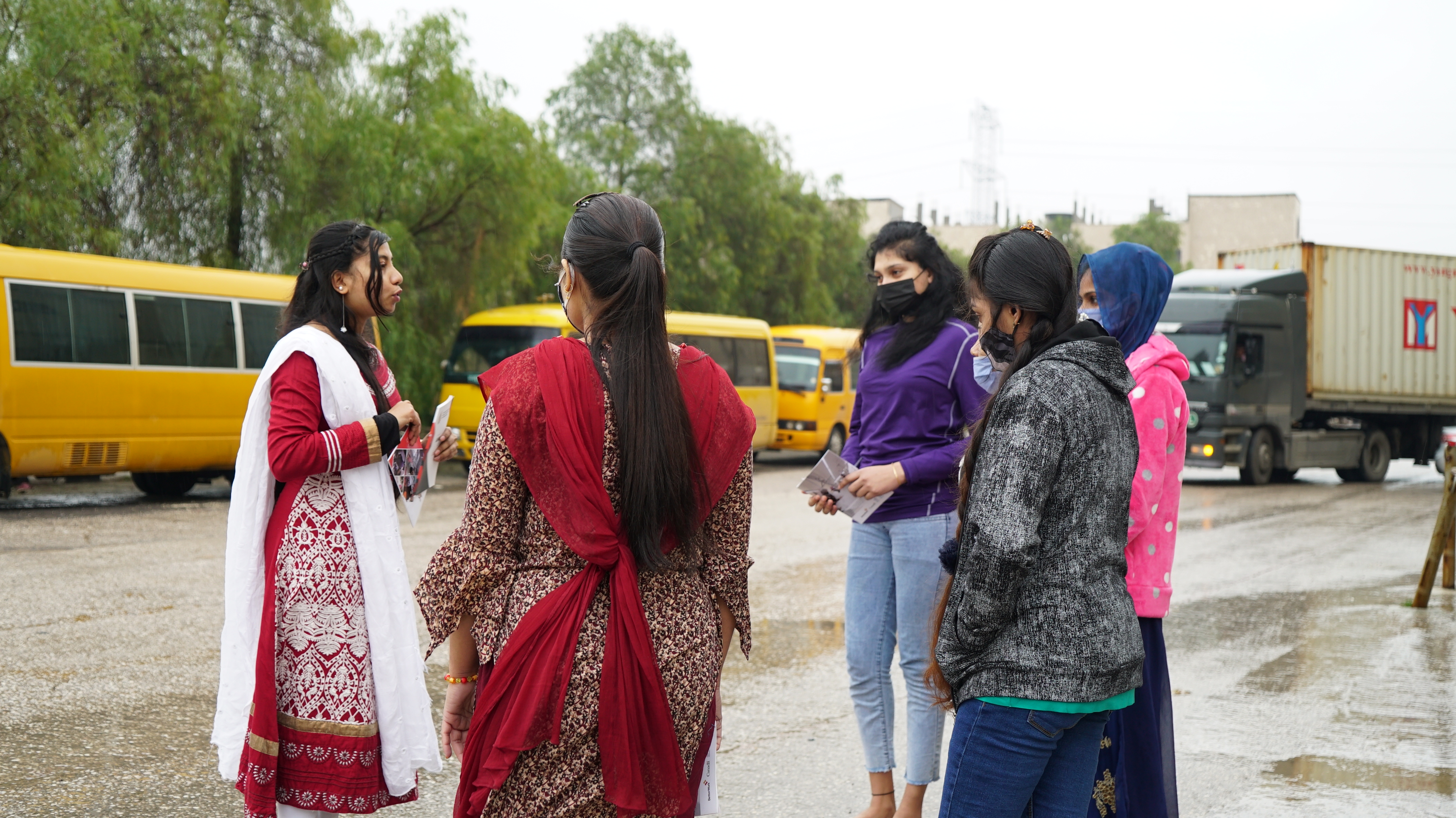 Maya talks to other migrant garment workers, all young women, outside by a road. A truck and buses are in the background.