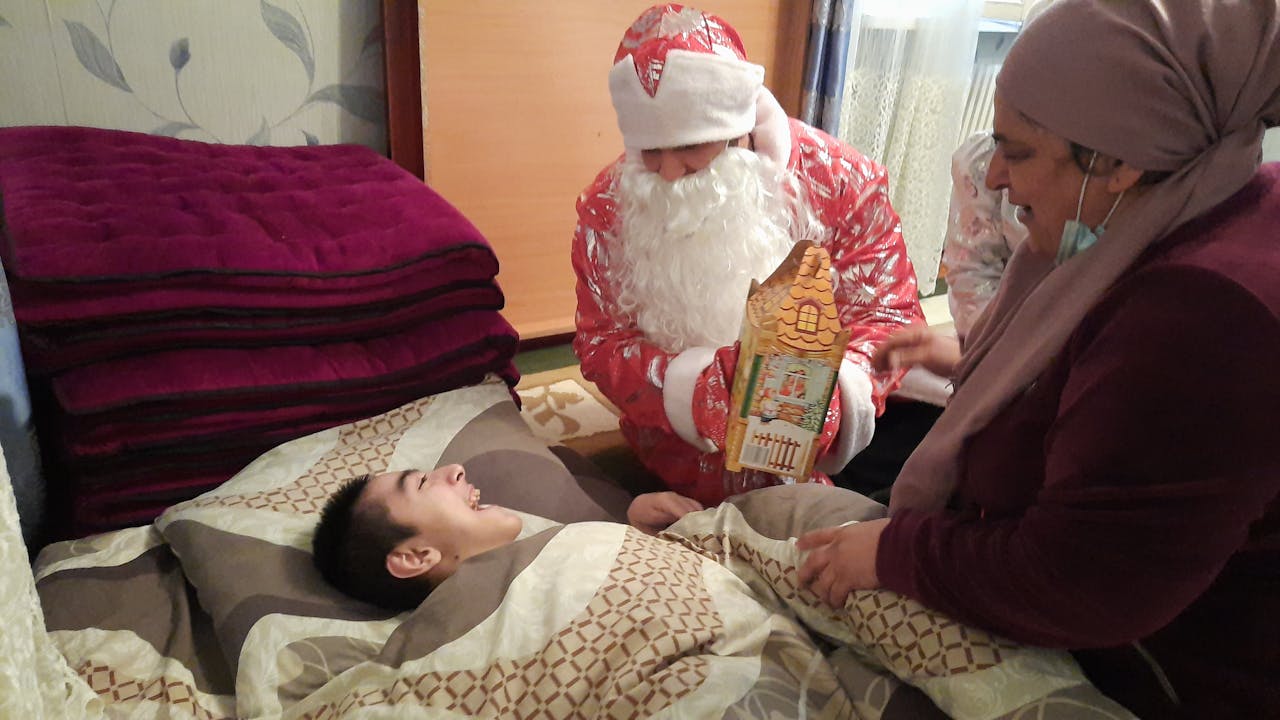 Une personne déguisée en Père Noël tient un cadeau et se tient à côté d'un lit. Dans le lit se trouve un petit enfant handicapé. L'enfant fait un grand sourire au Père Noël. Une femme portant un foulard les regarde.