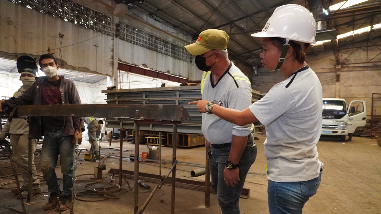 Myrna Pitaluna-Alngog speaks to three male colleagues in a warehouse and points at a task that needs to be done. She wears a hard hat, a t-shirt and jeans.
