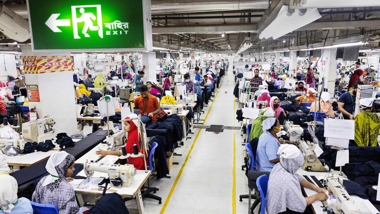 A wide shot of the ready-made garment factory floor. Rows of people operate sewing machines. In the forefront is a green emergency exit sign.