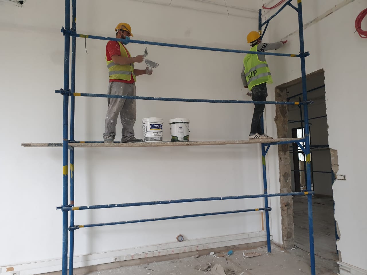 Dos hombres se suben a un andamio en una habitación y pintan una pared de blanco.